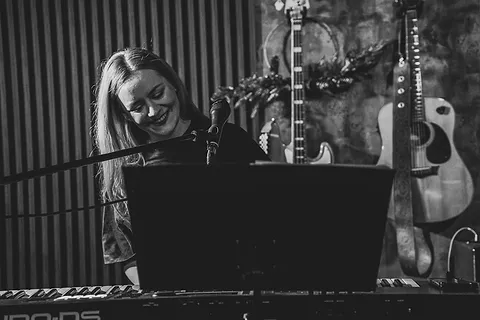 Uśmiechnięta wokalistka siedzi przy keyboardzie, w tle gitary akustyczne i świąteczna dekoracja, czarno-białe zdjęcie z koncertu