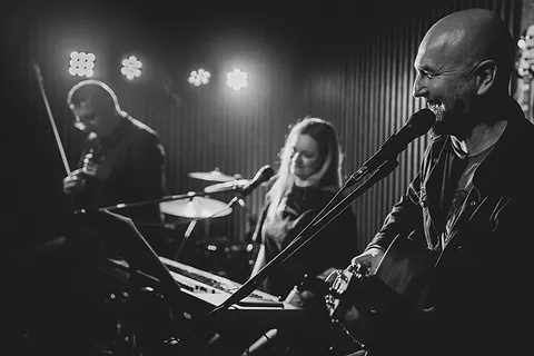 Wokalista z gitarą w pierwszym planie, za nim wokalistka przy perkusji i skrzypek na scenie, czarno-białe zdjęcie z koncertu