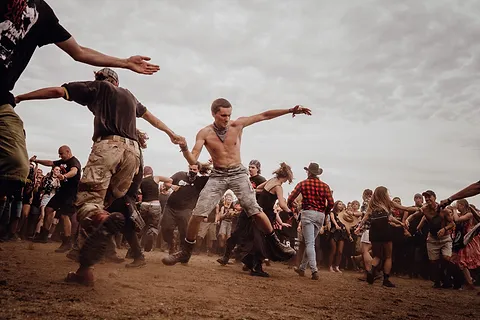 Pogo pod sceną – ludzie w skoku z rozpostartymi rękami na zakurzonym polu festiwalowym