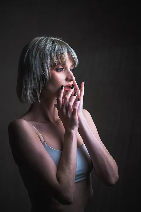 Portret kobiety z krótkimi jasnymi włosami i grzywką, dłonie przy twarzy, ciemne tło