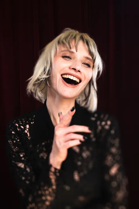 Roześmiana kobieta z blond fryzurą bob w czarnej koronkowej bluzce na tle czerwonej kurtyny