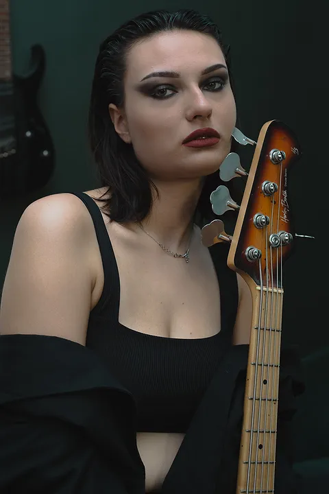 Młoda kobieta z mocnym makijażem oczu trzyma gitarę basową, w tle zielona ściana