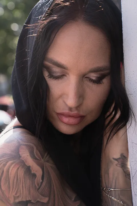 Portret kobiety w kapturze z opuszczonym wzrokiem, tatuaże na ramionach, światło dzienne